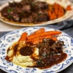 A blue and white plate with mashed potatoes and gravy, beef roast and gravy and roasted carrots.