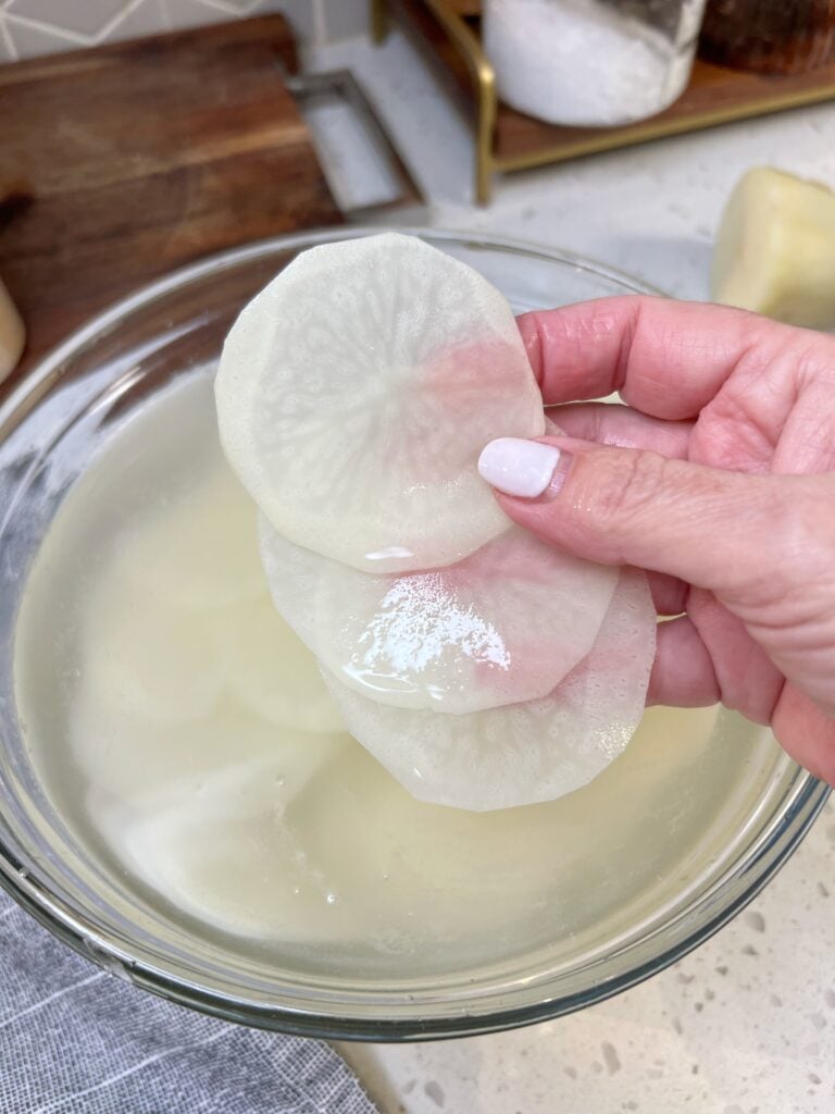 Me holding three thinly sliced potatoes over a bowl of water.