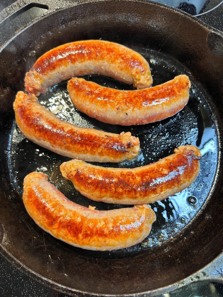 Five Italian hot sausages searing in a cast iron pan. The sausages are golden brown.