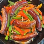 Five seared Italian hot sausages in a cast iron pan with thin strips of bell peppers in green, red and orange and thinly sliced onions.