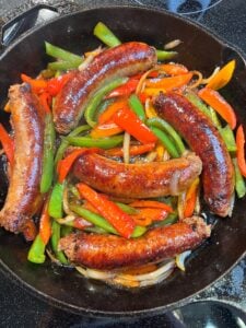Five seared Italian hot sausages in a cast iron pan with thin strips of bell peppers in green, red and orange and thinly sliced onions.