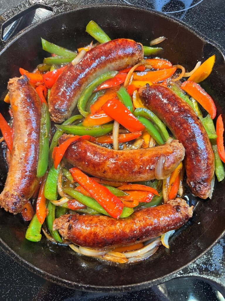 Five seared Italian hot sausages in a cast iron pan with thin strips of bell peppers in green, red and orange and thinly sliced onions.