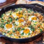 A cheesy breakfast skillet in a cast iron pan. The skillet has ground breakfast sausage, diced potatoes, and spinach. It's topped with cheese and five sunny side up eggs.