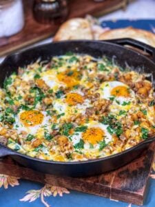 A cheesy breakfast skillet in a cast iron pan. The skillet has ground breakfast sausage, diced potatoes, and spinach. It's topped with cheese and five sunny side up eggs.
