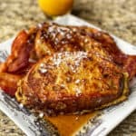 Three pieces of the best French toast on a square white platter topped with syrup and powdered sugar.