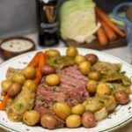 A platter of traditional corned beef and cabbage. The sliced corned beef is running down the center with cooked carrots on one side and cooked cabbage and baby potatoes scattered around the corned beef. The dish is garnished with fresh chopped parsley.