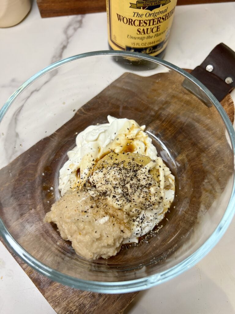 Ingredients in a small glass bowl for creamy horseradish sauce to pair with traditional corned beef and cabbage. The ingredients include sour cream, salt, pepper, Worcestershire sauce, horseradish and Dijon mustard. They are not mixed together.