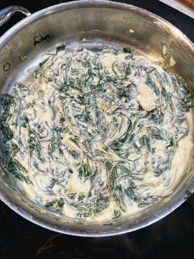 Cream cheese melted with baby spinach in a stainless steel pan for six cheese spinach artichoke dip.