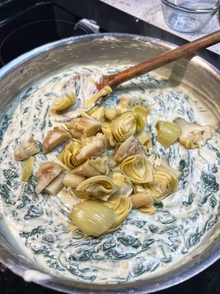 Chopped artichokes added to a creamy, cheesy spinach mixture in a stainless steel pan.