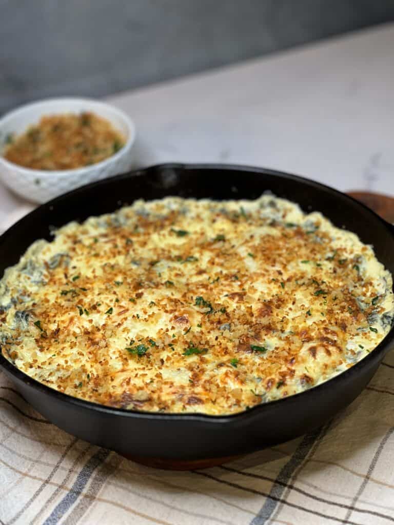 A cast iron pan filled with baked, six cheese spinach artichoke dip that has been sprinkled with toasted breadcrumbs.