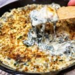 A cast iron pan filled with baked, creamy six cheese spinach artichoke dip that has a toasty breadcrumb topping. I am holding a long cracker that has been dipped in the middle and has the dip on top of the cracker and the cheese is pulling down.