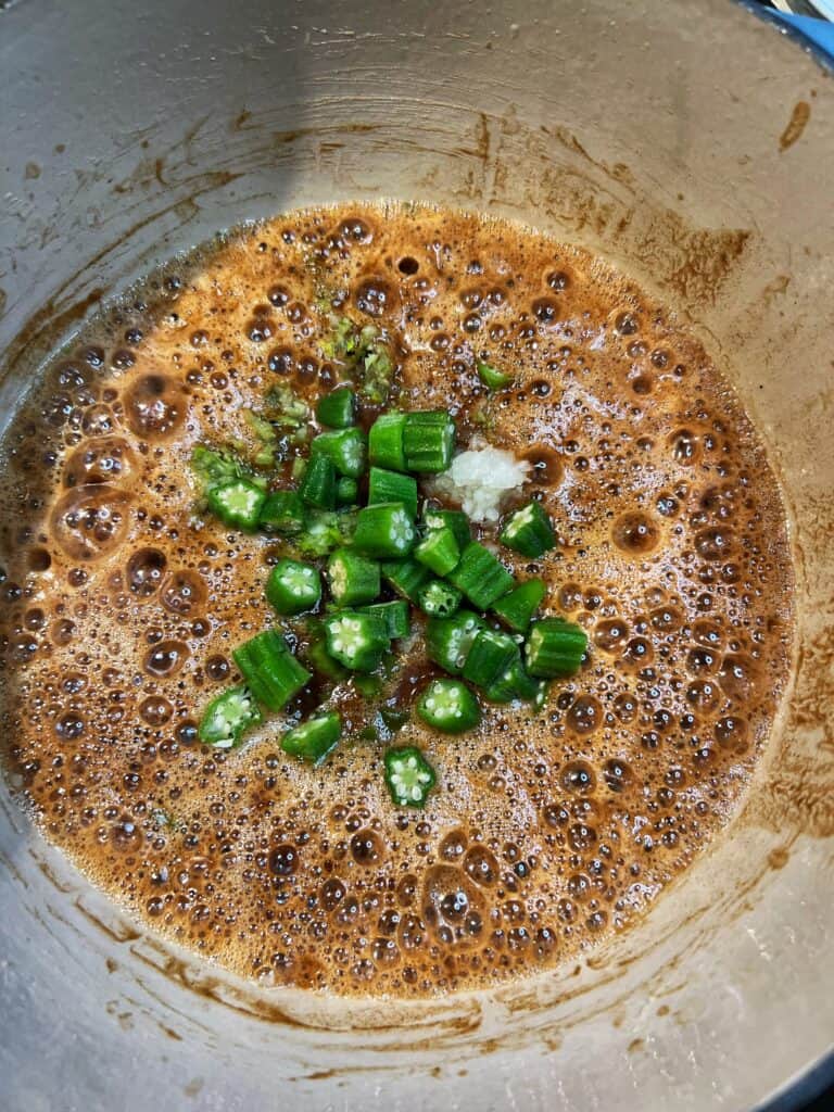 Diced celery, bell pepper, onions and okra added to a pot of cooked roux. The roux is a dark brown. 