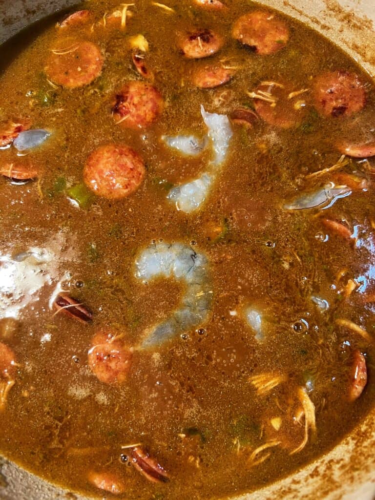 Raw shrimp added to a pot of gumbo. The gumbo is a deep dark brown color.