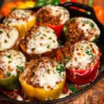 Eight baked sausage stuffed bell peppers in a variety of colors in a cast iron pan. Each pepper has melty mozzarella cheese on top and is garnished with fresh chopped parsley.