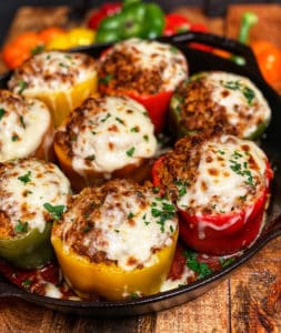 Eight baked sausage stuffed bell peppers in a variety of colors in a cast iron pan. Each pepper has melty mozzarella cheese on top and is garnished with fresh chopped parsley.