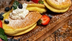 three Japanese soufflé pancakes on a wooden board topped with strawberries, blueberries, powdered sugar and whipped cream