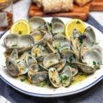 A large white bowl of butter garlic steamed clams. The clams are open and in a broth with fresh chopped parsley and lemon wedges.