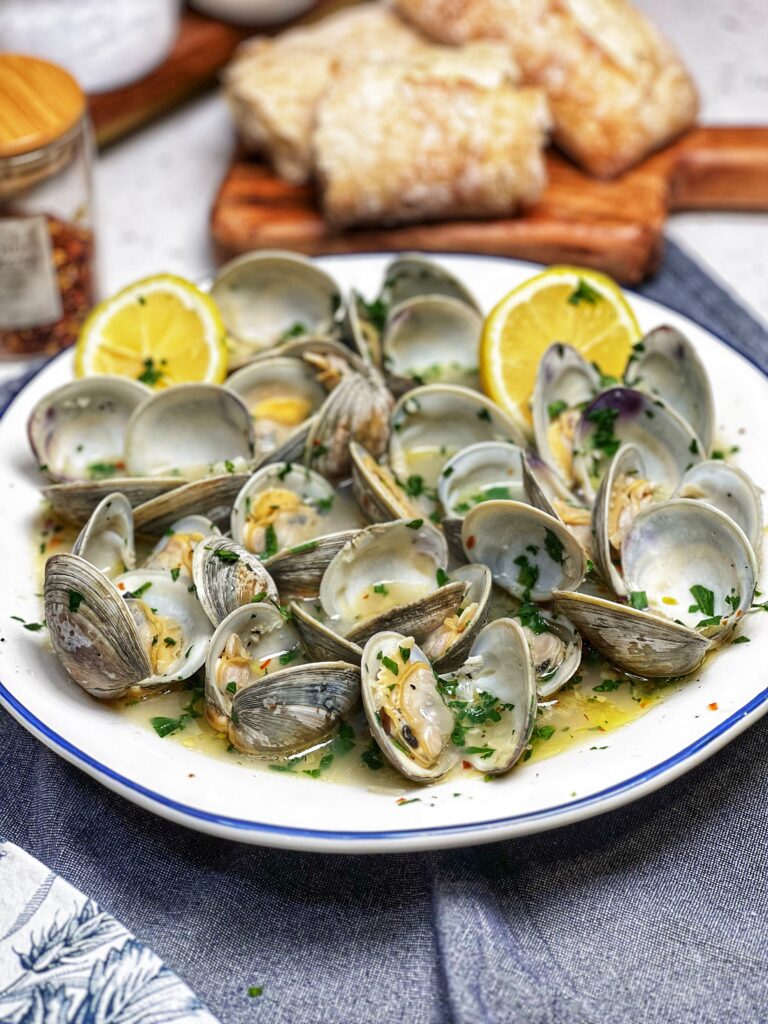 A large white bowl of butter garlic steamed clams. The clams are open and in a broth with fresh chopped parsley and lemon wedges.