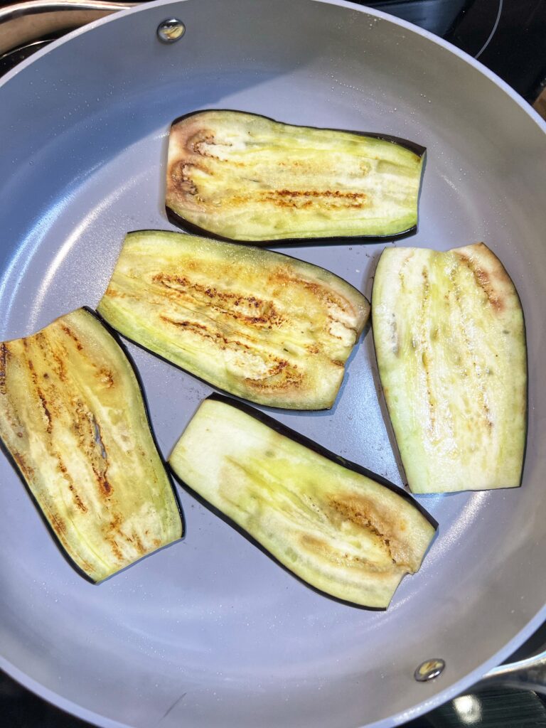 Five slices of eggplant sautéing in a non-stick pan.