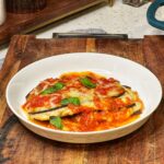 A white dish holding a stack of low carb eggplant Parmesan. Three layers of sautéed eggplant with ricotta cheese in the middle and finished with marinara and melty cheese.