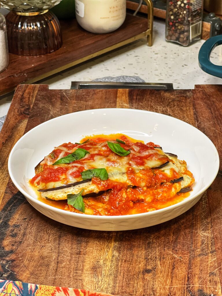A white dish holding a stack of low carb eggplant Parmesan. Three layers of sautéed eggplant with ricotta cheese in the middle and finished with marinara and melty cheese.