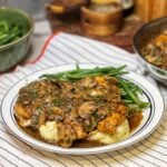 A white plate trimmed in blue stripes holding steamed green beans and mashed potatoes that are topped with two pieces of chicken marsala. The dish is garnished with fresh parsley.