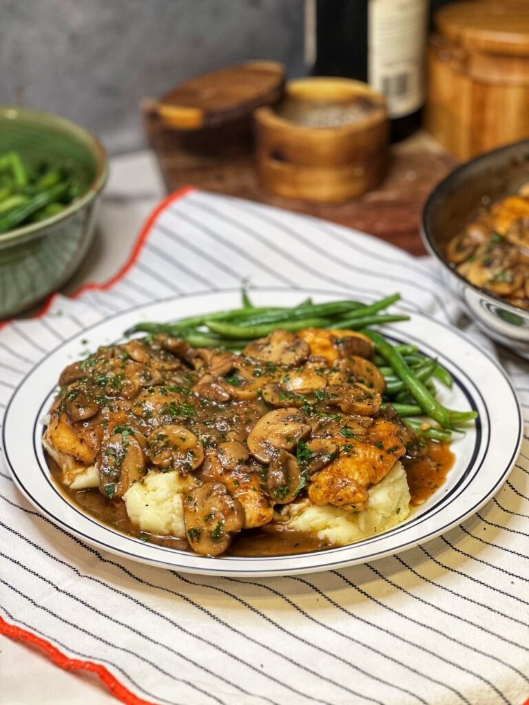 A white plate trimmed in blue stripes holding steamed green beans and mashed potatoes that are topped with two pieces of chicken marsala. The dish is garnished with fresh parsley.