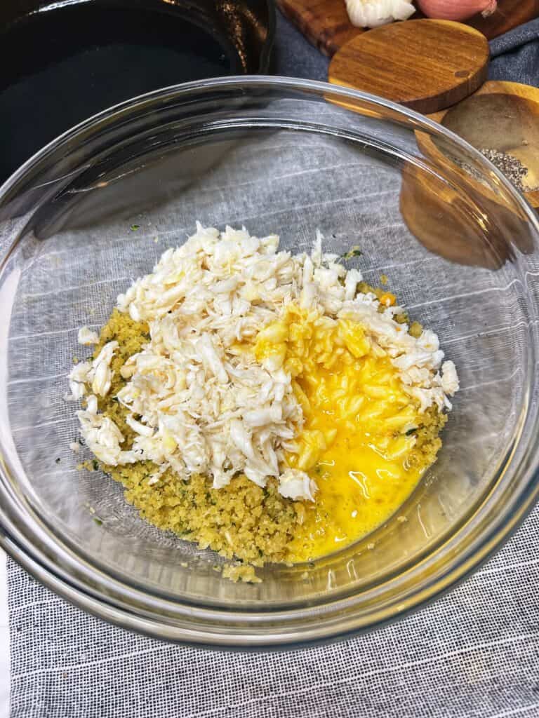 A glass bowl filled with a breadcrumb mixture, crabmeat and a beaten egg.