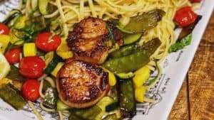 a white plate of seared scallops with roasted summer vegetables