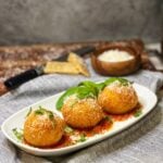 Three arancini Italian rice balls sitting on a long platter. Each are sitting on a dollop of marinara and garnished with freshly grated Parmesan cheese and fresh basil.