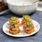 A small white plate holding four spicy tuna rice squares. The rice squares are crispy and browned. They are topping with a fresh tuna mixture and topped with black sesame seeds and a slice of jalapeño.