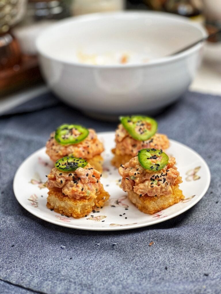 A small white plate holding four spicy tuna rice squares. The rice squares are crispy and browned. They are topping with a fresh tuna mixture and topped with black sesame seeds and a slice of jalapeño.
