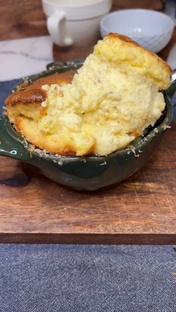 Showing a spoonful of a fluffy cheesy soufflé that has been baked in a round, green cocotte.