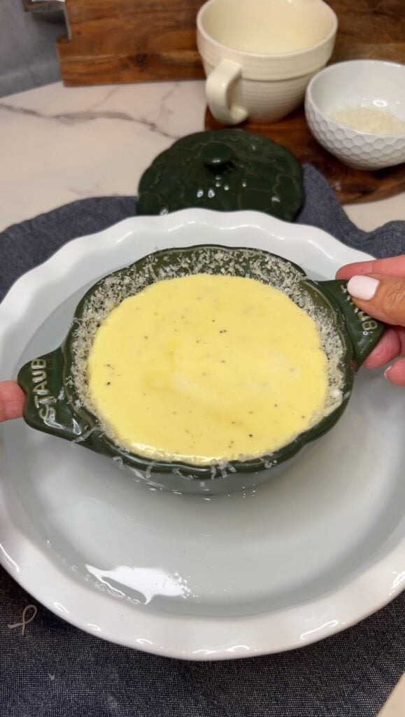 A found, green cocotte filled with a creamy cheesy egg mixture for a soufflé. The cocotte is being lowered into a round, white pie dish that is filled with water.