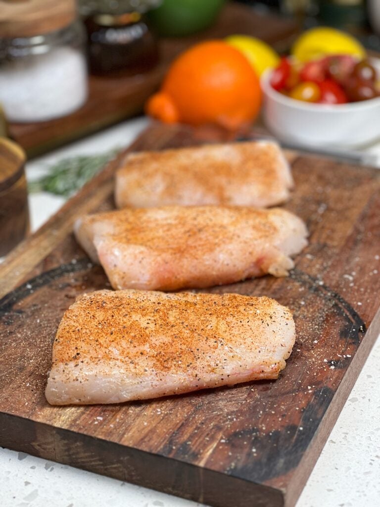 Three pieces of raw corvina fish seasoned with salt, pepper and paprika. The fish is sitting on a wooden cutting board.