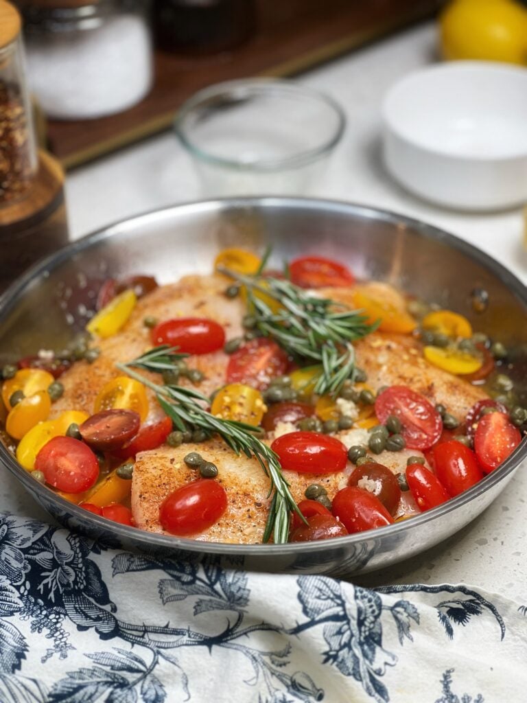 Three pieces of seasoned corvina fish in a stainless steel pan with halved cherry tomatoes, rosemary sprigs, garlic, lemon juice and orange juice.