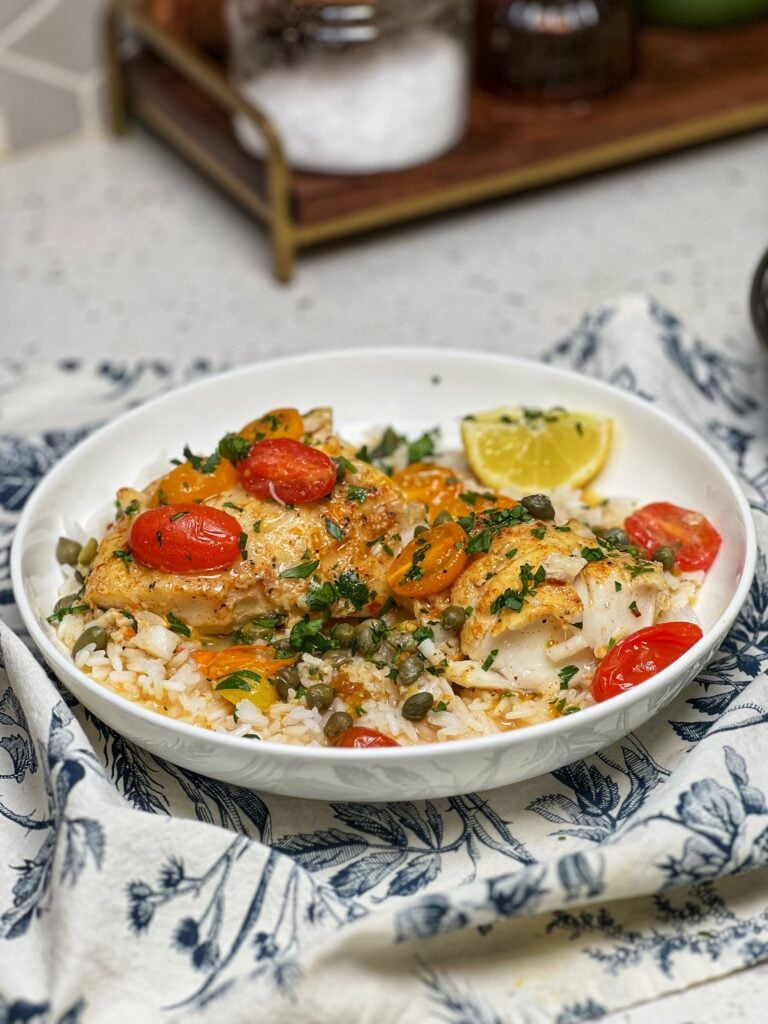  A white bowl of baked corvina with tomatoes. The fish is over a bed of rice with cherry tomatoes and garnished with fresh parsley.