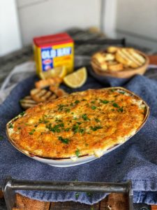 an oven-safe dish filled with hot crab dip with a container of Old Bay seasoning in the background and crostini