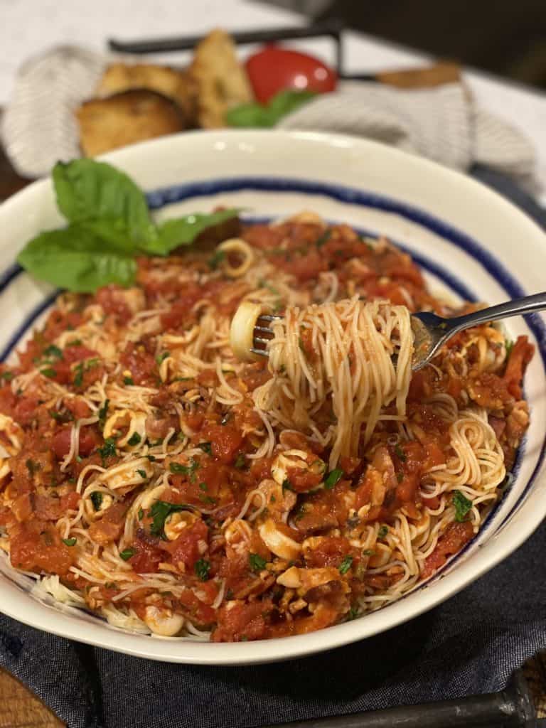 a big white bowl with blue stripes filled with spicy calamari pasta garnished with basil leaves and fresh parsley and a fork twirled with the pasta