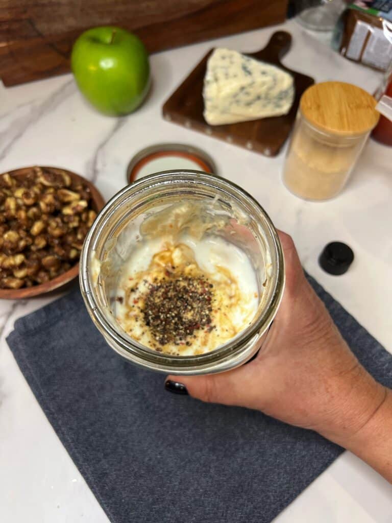 Ingredients for a Red Roquefort dressing in a large glass jar. The contents include, mayo, buttermilk, garlic powder, ketchup and black pepper. I am holding the jar.