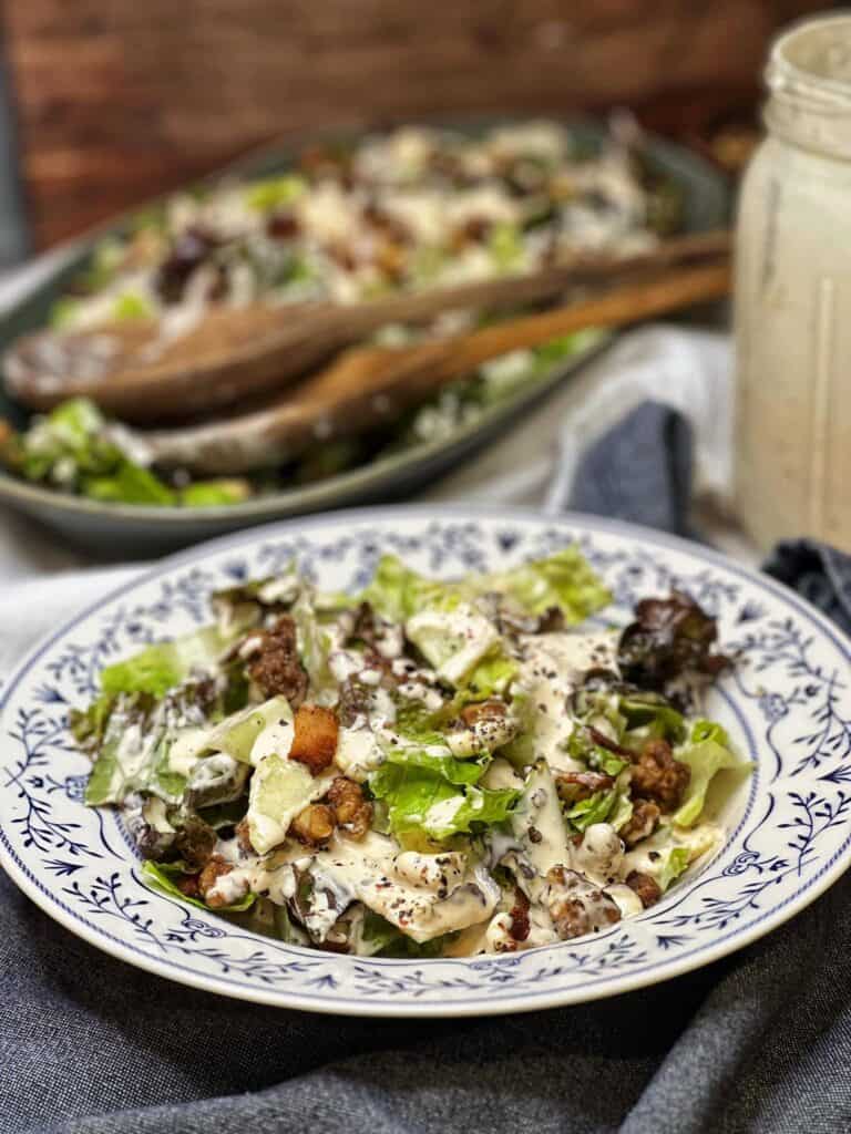 A blue flowered plated holding a serving of a Red Roquefort Salad. The salad includes chopped Romaine, chopped red lettuce, covered in a creamy white dressing and topped with crisp bacon and candied walnuts.