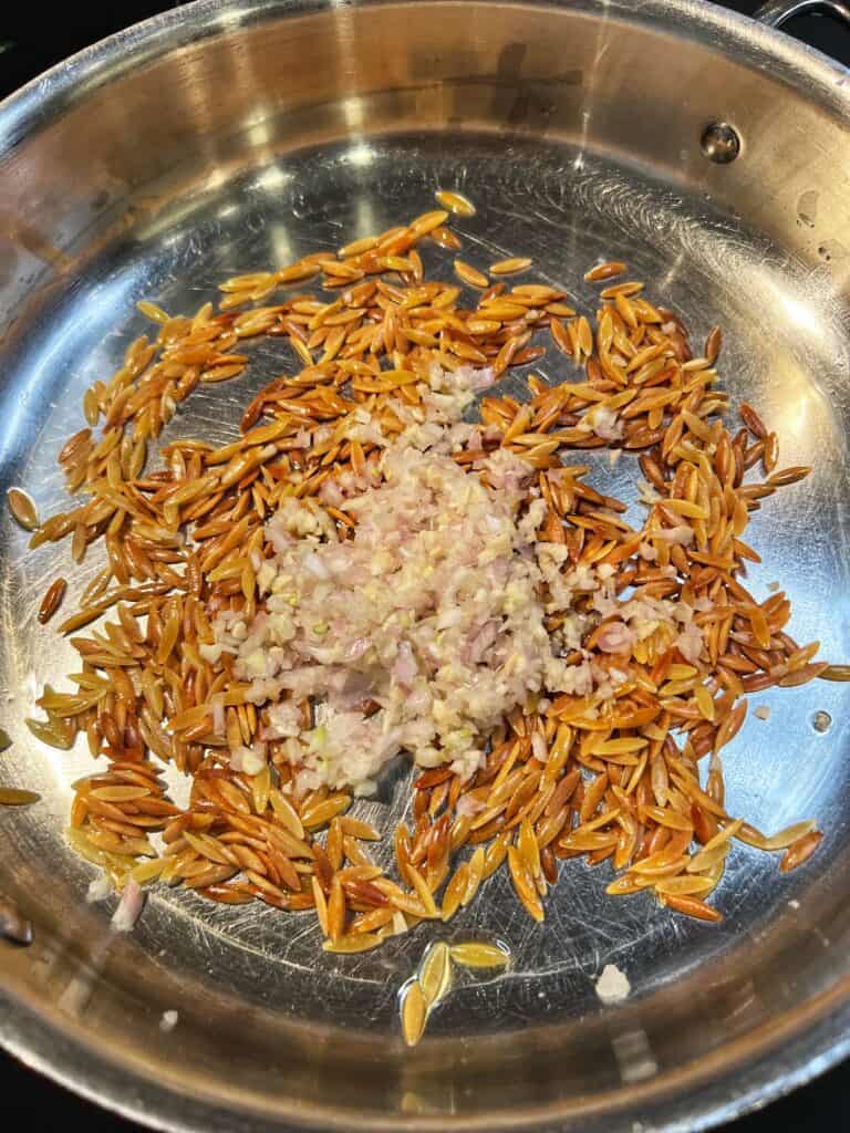 Browned orzo in a stainless steel pan with minced garlic and shallots.