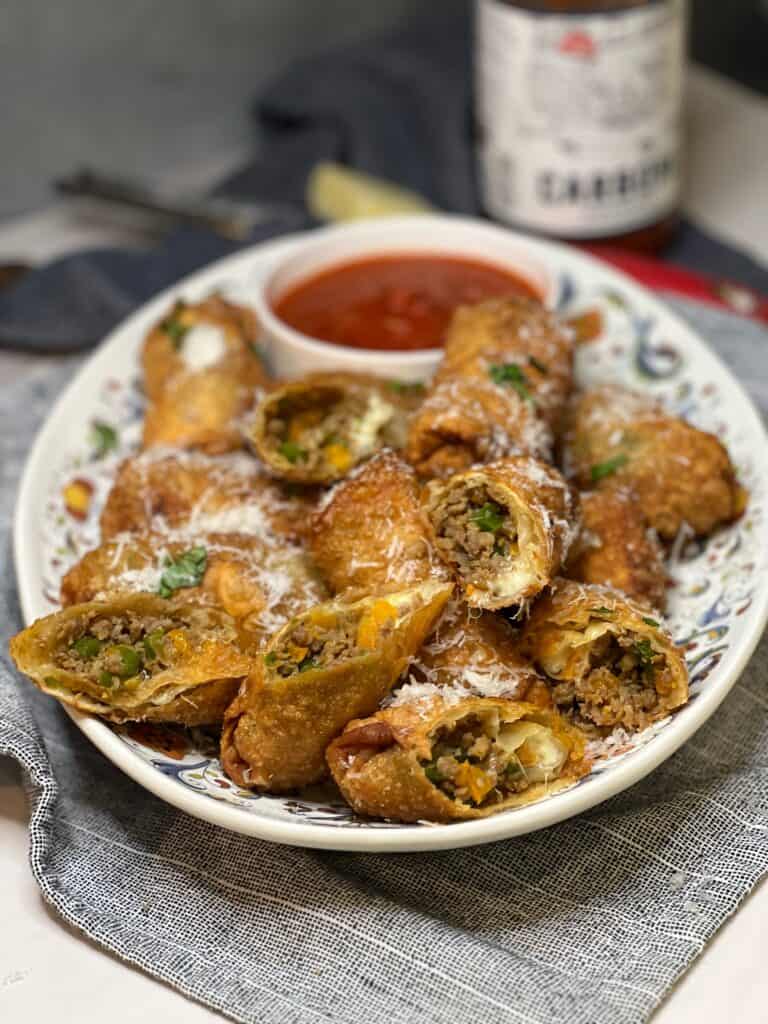 A platter of sausage and peppers egg rolls with marinara sauce in a small bowl. Some of the egg rolls are opened to see the inside. The egg rolls are garnished with fresh basil and freshly grated Parmesan cheese.