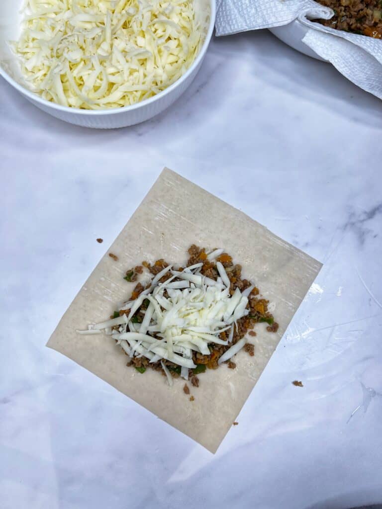 Sausage and peppers and shredded mozzarella cheese on a corner of an egg roll wrapper.
