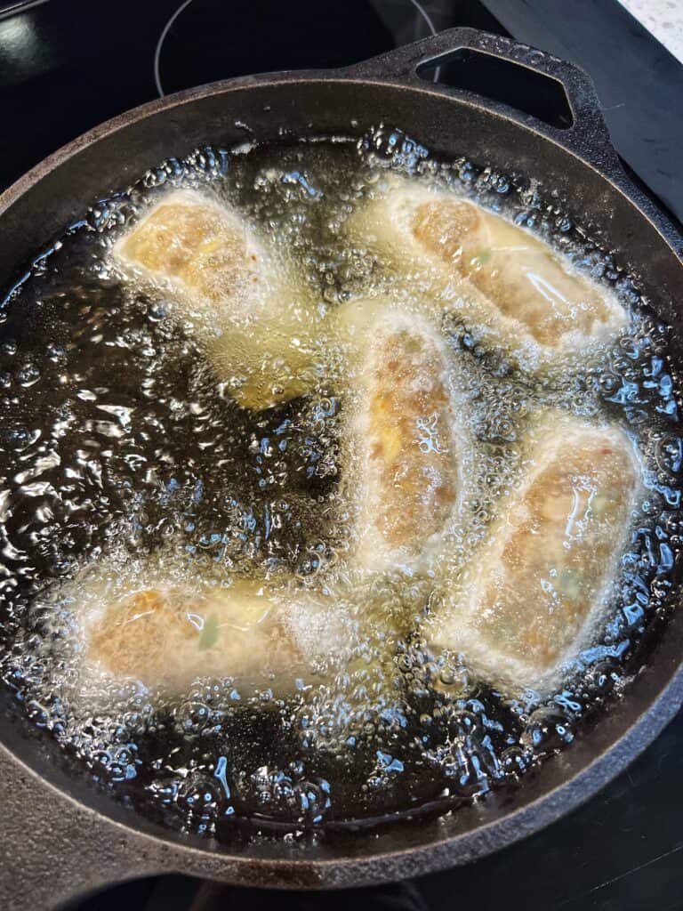 Five egg rolls frying in a deep cast iron pan.
