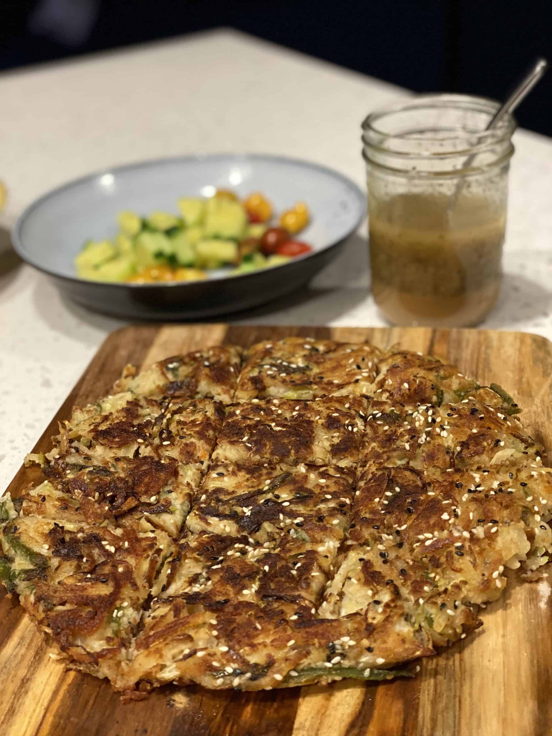 A vegetarian jeon (pancake) on a wooden board with a bowl of salad in the background