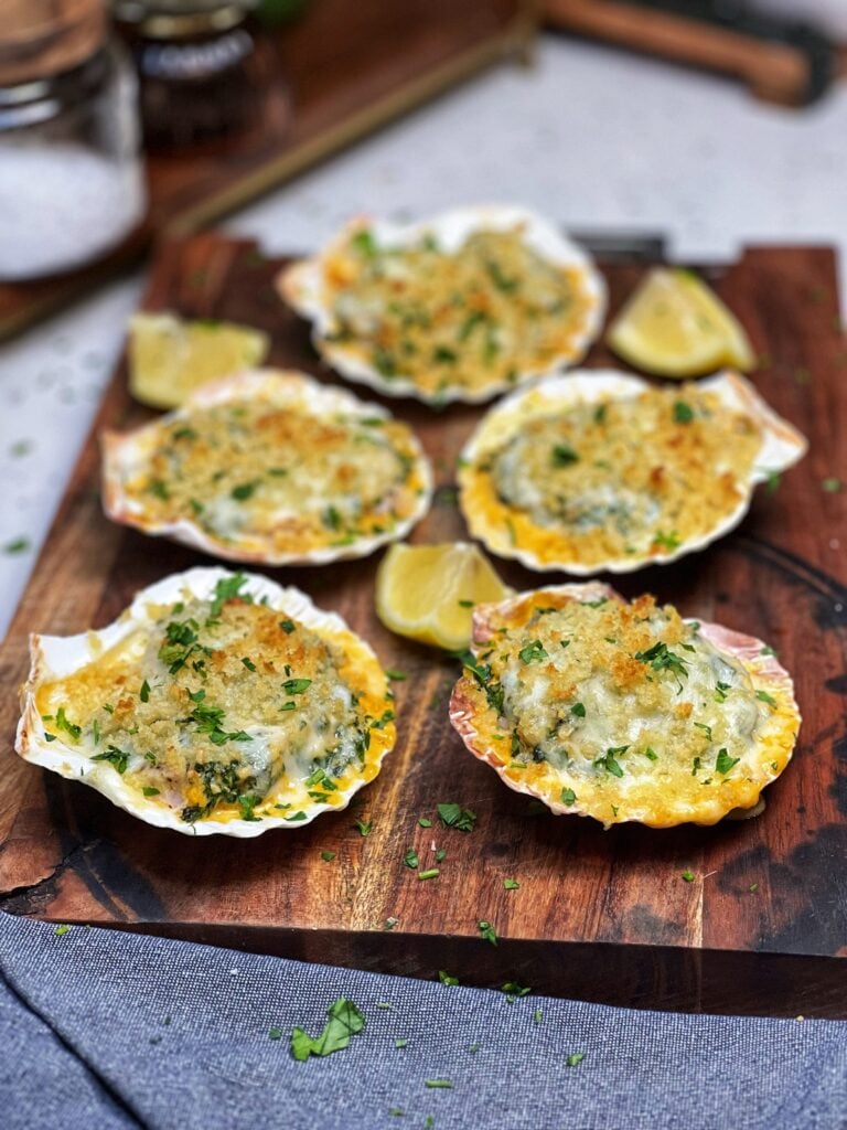 Five shrimp Rockefeller sitting on a wooden board. There are five scallop shells filled with the Rockefeller mixture which is golden brown on the top.