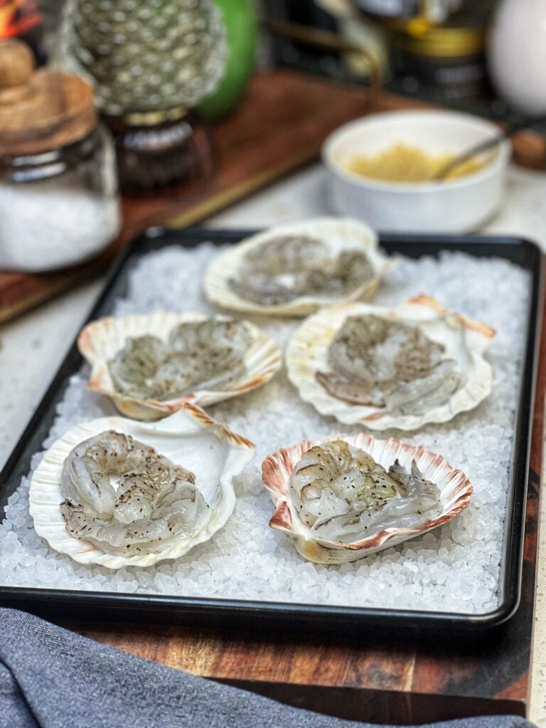 Shrimp seasoned with salt and pepper laying inside 5 scallop shells. The shells are sitting on a sheet pan that has been filled with rock salt.