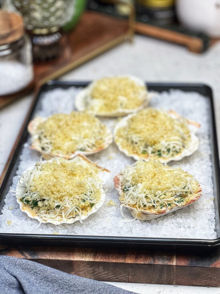 Five scallop shells sitting in a pan lined with rock salt. Each shell is filled with shrimp, a creamy sauce, cheese and a buttery panko topping.