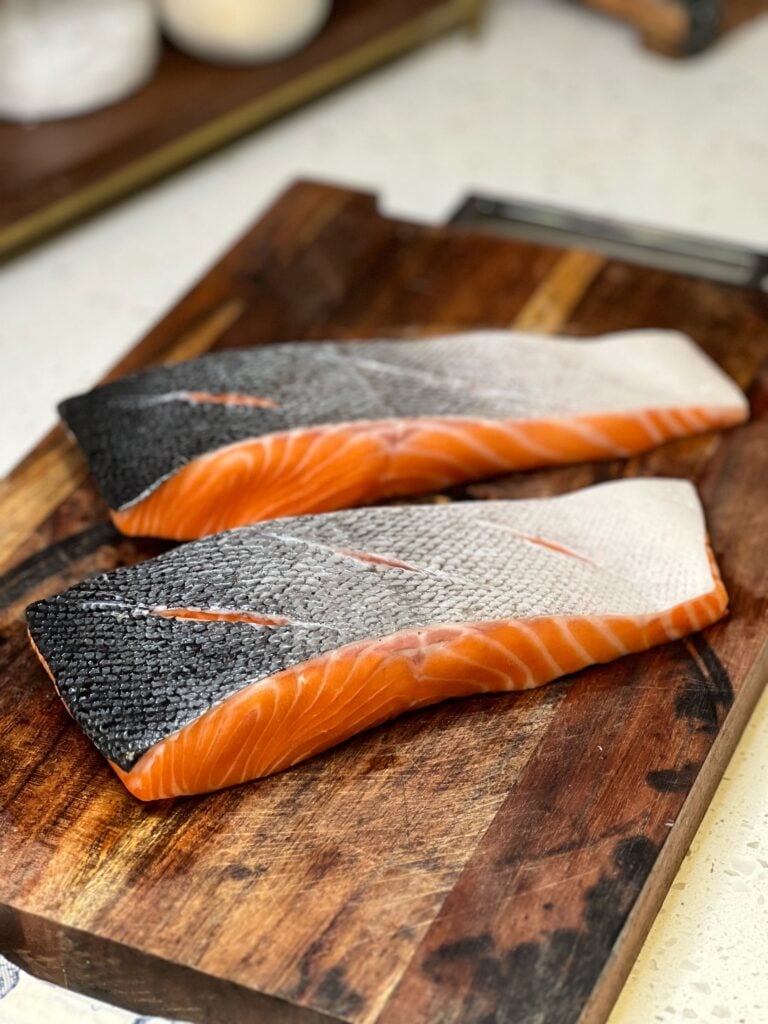 Two pieces of salmon sitting on a wooden cutting board. The salmon is skin-side up and each piece has three scores in the skin.